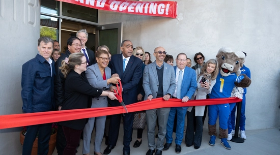 Approximately 20 people lined up for a ribbon cutting ceremony under a banner that reads "Grand Opening." Los Angeles Mayor Karen Bass is holding a large pair of red scissors next to a red ribbon.