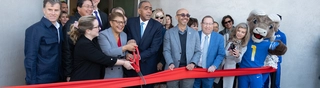Approximately 20 people lined up for a ribbon cutting ceremony under a banner that reads "Grand Opening." Los Angeles Mayor Karen Bass is holding a large pair of red scissors next to a red ribbon.